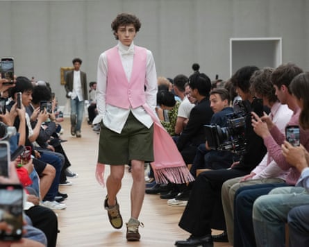 a male model wears a white dinner shirt with white bow tie, khaki shorts and a pale pink waistcoat with matching billowing scarf