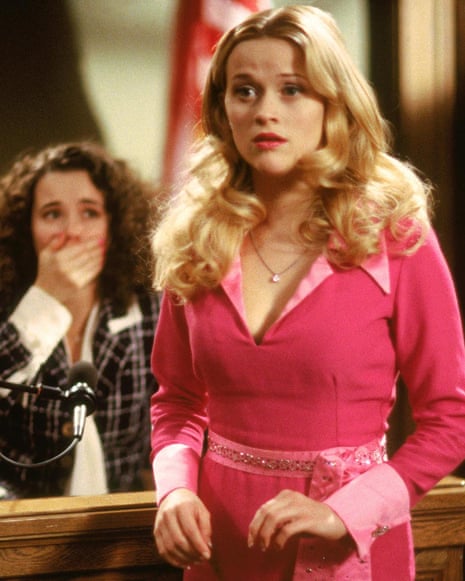 Reese Witherspoon in a bright pink suit stands at a wooden podium with another woman behind her covering her mouth