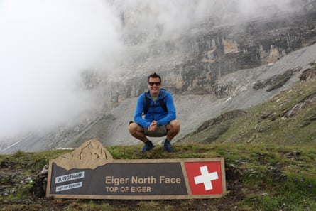 A man wearing a blue fleece and shorts, crouching by a sign saying Eiger North Face, with cloud behind him