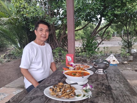 Man in a white T-shirt sits at a wooden table outside. Table has several dishes of food on it.