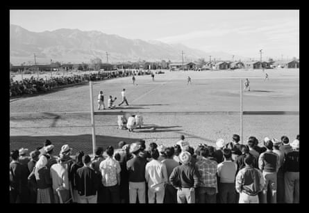 A crowd of mostly men standing (no bleachers) along the sidelines of a baseball game in a grassless field, with barracks and mountains beyond.
