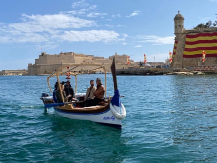 A group of people on an ancient looking boat on a deep blue sea on a beautiful sunny day with Malt Grand Harbour behind them