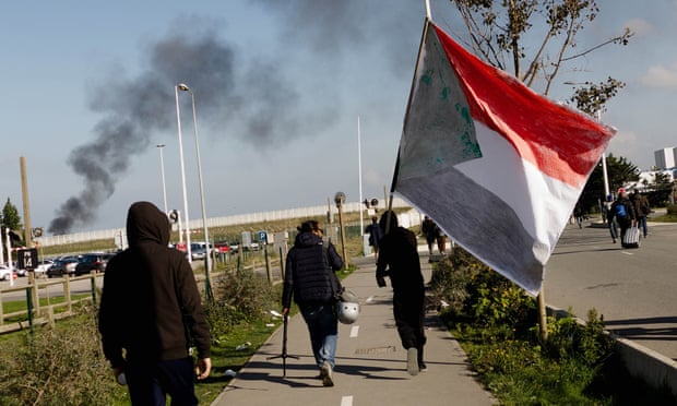 Migrants leave the refugee camp in Calais last October(one carrying the Sudanese flag).