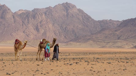Two figures crossing a mountainous desert with camels