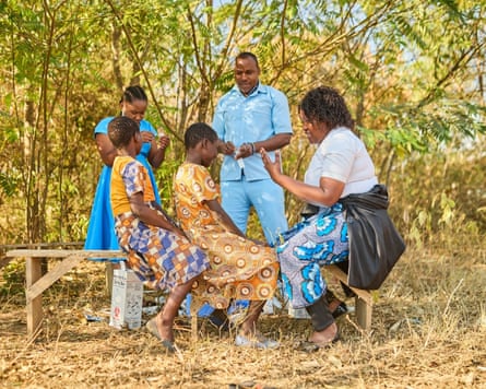 Two girls sit astride a bench in a leafy setting facing a woman who is also sitting on the bench. A girl and a man stand next to them