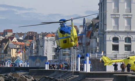An emergency helicopter takes off watched by onlookers and with another helicopter on the ground