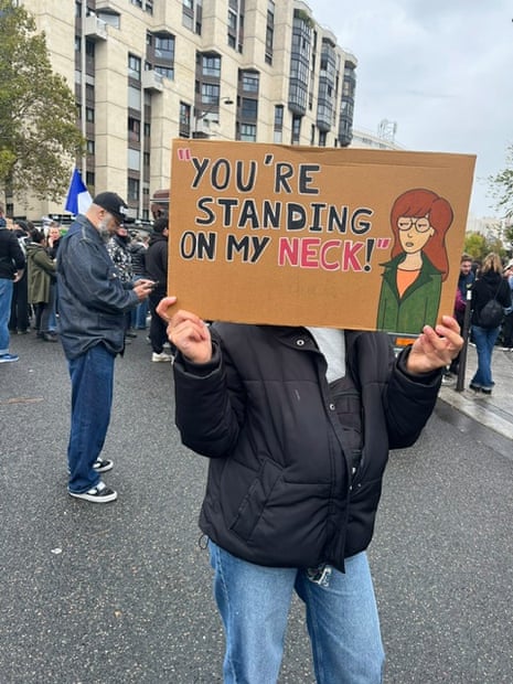 A protester in the Paris march on 24 October 2026 holding a protest sign featuring cartoon figure Daria with the message 'You're standing on my neck'