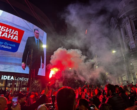 Crowd in Belgrade holds flares releasing red smoke near a screen showing Serbia's ousted leader