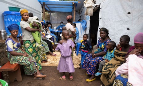 Women and children sit on benches or stand in a makeshift waiting area outside a tent.