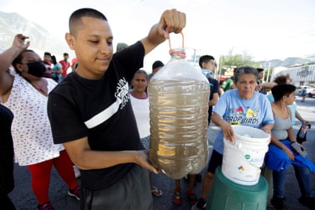 A man holds up a large water bottle of muddy brown water as people queue with buckets to collect water