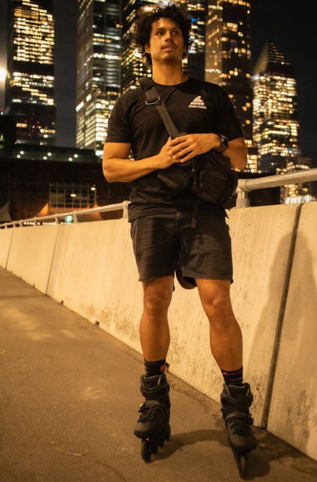 Miguel Ramirez, in T-shirt and shorts, wearing his skates on a path with high-rise office blocks behind at night