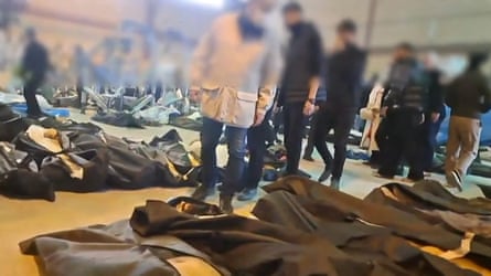 People walk among rows of dead people wrapped in black sheets or blankets.