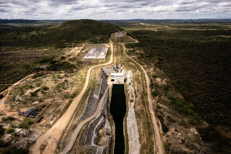 Aerial view of a canal dug into semi-arid land with a building at the top of the channel and electrical transformers and cables on steel lattice towers structures