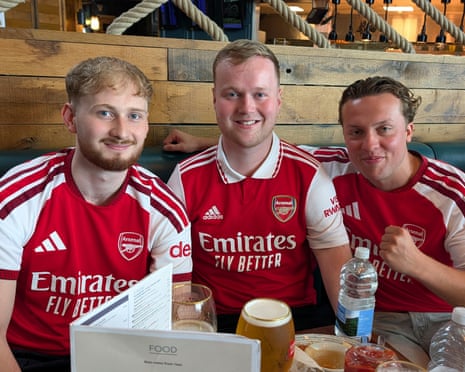 Three young men wearing Arsenal football shirts sitting at a pub table with beer and water in front of them