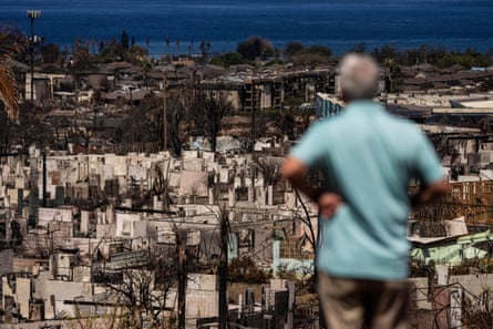 An older man, seemingly white, with white hair and wearing a teal polo shirt and khakis, is blurred in the right foreground as he looks far downward onto the burned remains of an oceanside town.