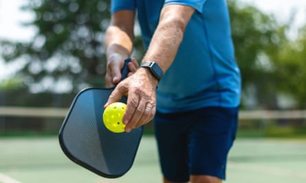 A man on an outdoor court holds a pickleball racket and ball 