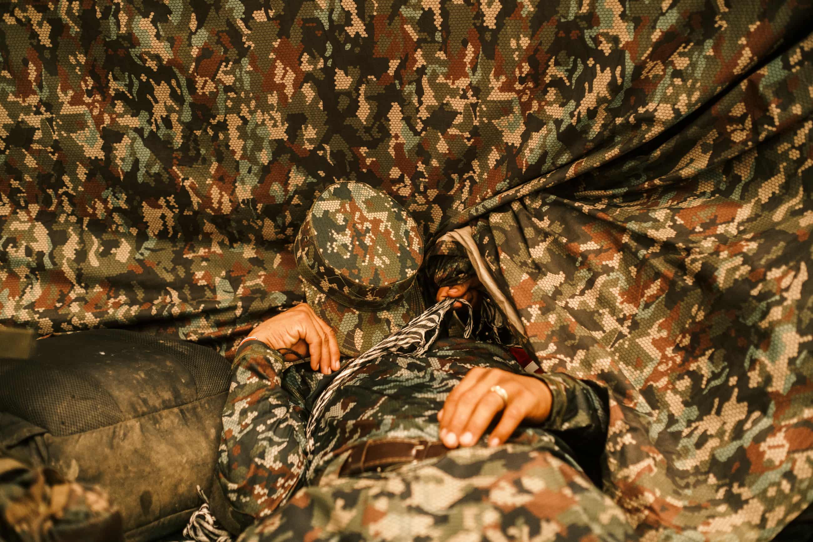 A fighter in camouflage gear asleep with their cap over their face in a tent of the same material