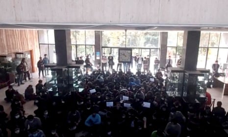 A crowd of people holding placards sit in a building's large sunlit entrance hall
