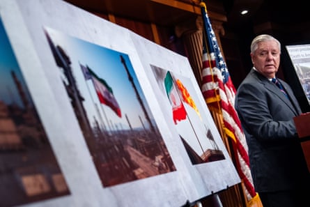 A man stands in a oom with a US flag in front of posters with photographs showing the Iranian flag