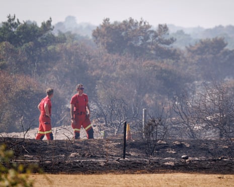 Two firefighters dressed in red on scorched land with smoke in distance.
