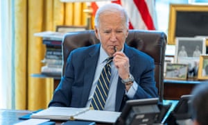 Jo Biden at desk sucking a pen with US flag and framed photos behind him
