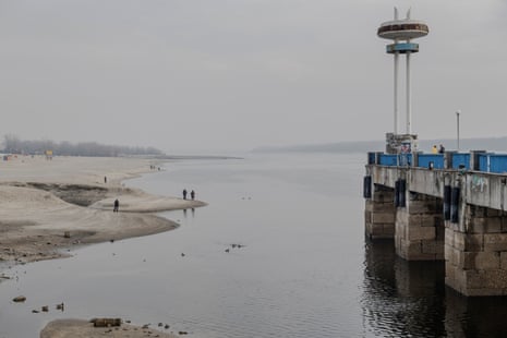 People walk along the shore of a river with a concrete structure in the water