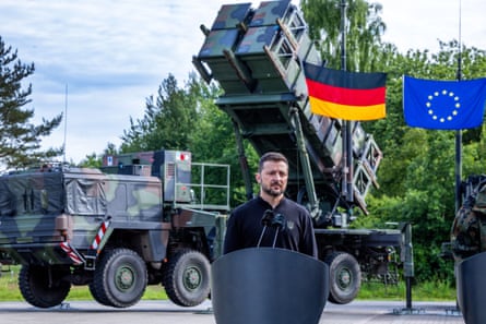 Zelenskyy stands at a small lectern with the Patriot system behind him
