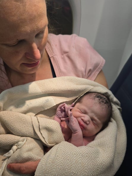 A mother in a pink shirt leans against a plane window while cradling her newborn baby, wrapped in an airline blanket