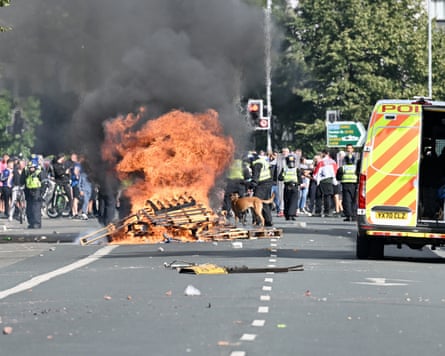 A bonfire burns in the road by a police van while officers attempt to control a crowd.