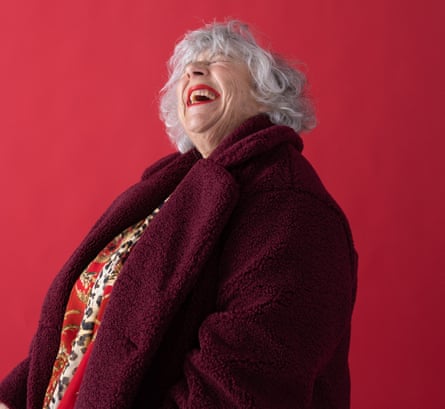 Actor Miriam Margolyes, head thrown back, laughing, wearing red patterned dress and burgundy coat, against red background