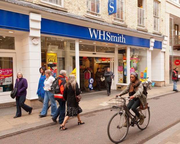 A woman cycling and five people walking past a WH Smith on a high street