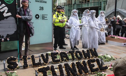 Black flowers are arranged to spell out the words Silvertown Tunnel in a funeral-style wreath as a group of protesters stand behind, wearing white robes, masks and headdresses