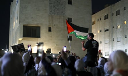 Demonstrators in a protest near the Israeli embassy in Amman, Jordan: a crowd of people is seen from behind, many holding up phone screens; one man is sitting on someone’s shoulder and a Palestinian flag is held aloft. It is dark and a tall building is illuminated in front of the crowd.