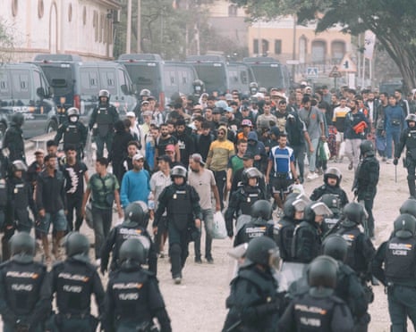 Dozens of young migrants walk escorted by riot police officers during the eviction of the makeshift camp on El Trampolin beach in Ceuta.