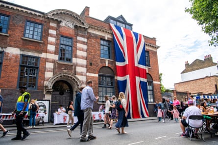 A huge union jack flag is draped over the side of a large red brick building as people sit at tables in the street and walk past.