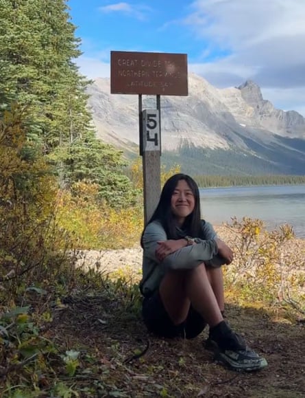 A woman sitting on the ground with her back against a trail marker, with dramatic mountains and a lake behind her.