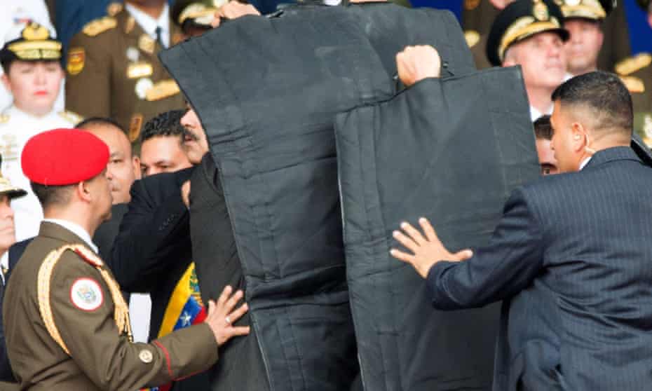 Security agents respond to a claimed assassination attempt on President Maduro, who is partly visible behind protective screen.