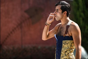 A man in matador trousers and braces but no top smokes a cigarette