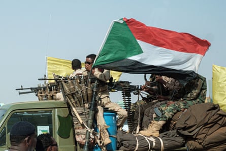 Armed fighters ride on a military vehicle with a Sudanese flag flying from it.