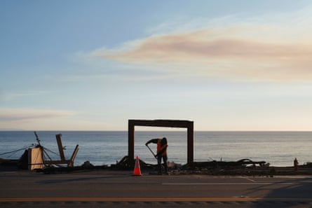 person with shovel stands framed by remains of a building, against the ocean in the background