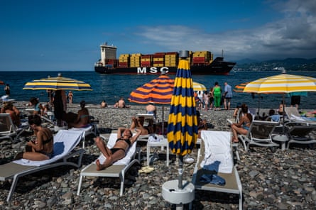 Beachgoers relax on loungers near Batumi as a large cargo ship passes behind striped umbrellas