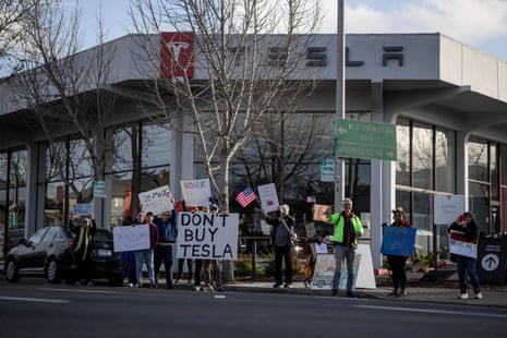 people hold signs against Tesla outside a dealership saying 'don't buy tesla' and 'de-musk america'