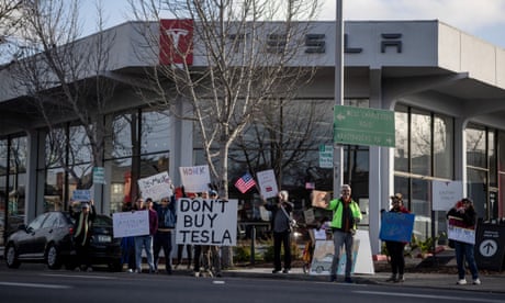 people hold signs against Tesla outside a dealership saying 'don't buy tesla' and 'de-musk america'