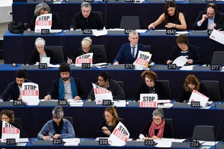People sitting at desks in rows hold up signs reading ‘Stop ISDS’.