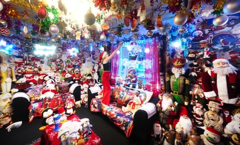 A girl stands on a sofa to hang a silver bauble from the ceiling of a room jam-packed with Christmas decorations