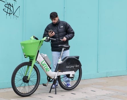 Sammy Gecsoyler looks at his phone while standing next to a Lime bike in front of a turquoise painted panelled wall