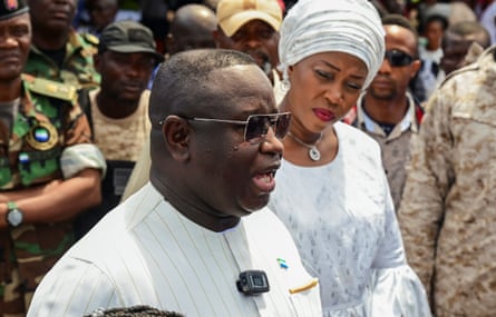 A middle-aged African man in a white shirt speaks to a crowd while a woman watches him
