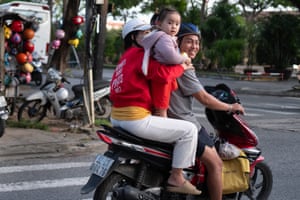 A child stands on the seat of a moped while an adult holds them. Another adult is holding the child.