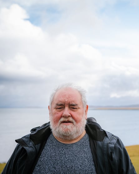 A heavy-set elderly man standing outside with a coastline view behind him