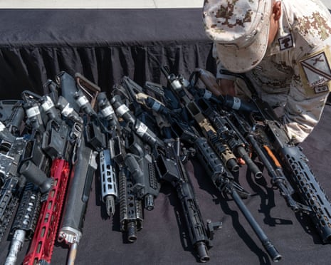 A soldier crouches to hold a rifle placed on a table alongside a number of other guns